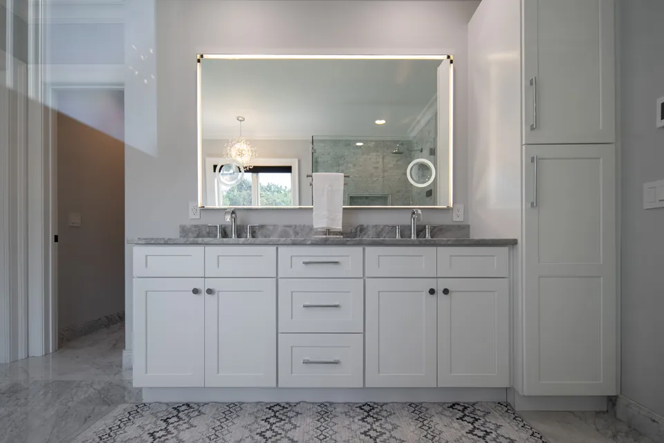 White double vanity with a gray stone top, backlit mirror and a tall linen cabinet on patterned floor tile