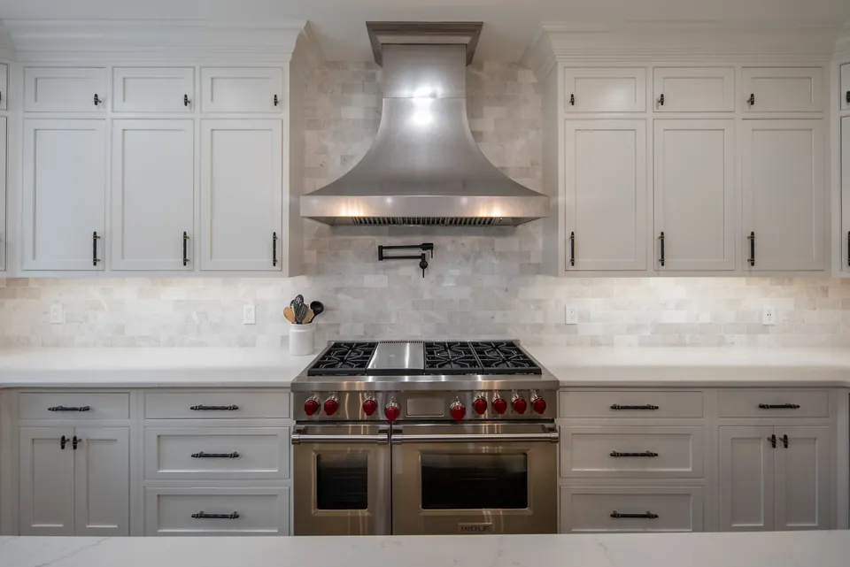 Stainless range hood over a dual-fuel range with a pot filler and marble subway tile backsplash between white cabinets