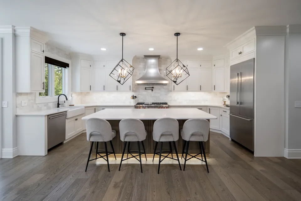 Remodeled white kitchen with a quartz island, four upholstered stools, geometric pendant lights and a Wolf range under a stainless hood