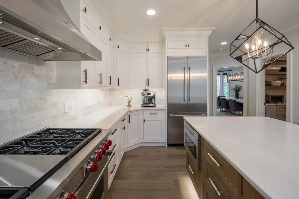 Professional range with red knobs, stainless hood and marble-look backsplash beside a built-in refrigerator