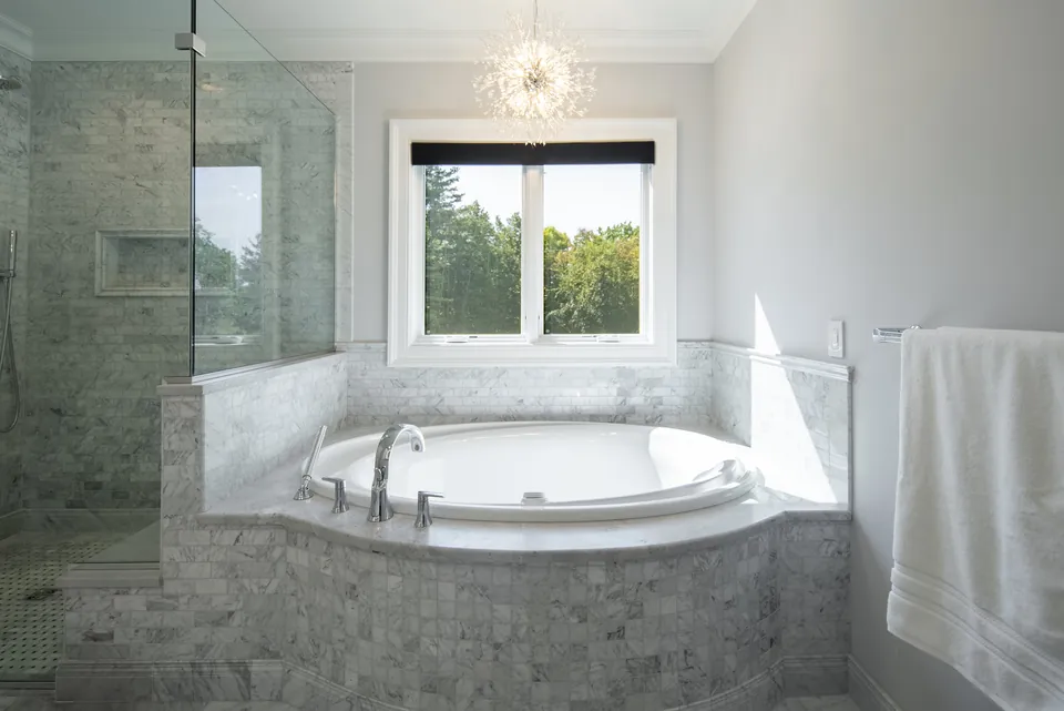 Soaking tub set in a marble mosaic tile deck under a window, next to a frameless glass shower