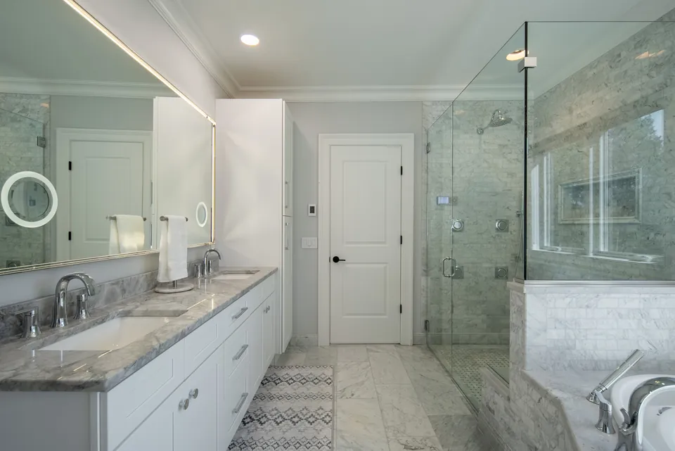 Double vanity with a backlit LED mirror facing a frameless glass shower lined with marble tile